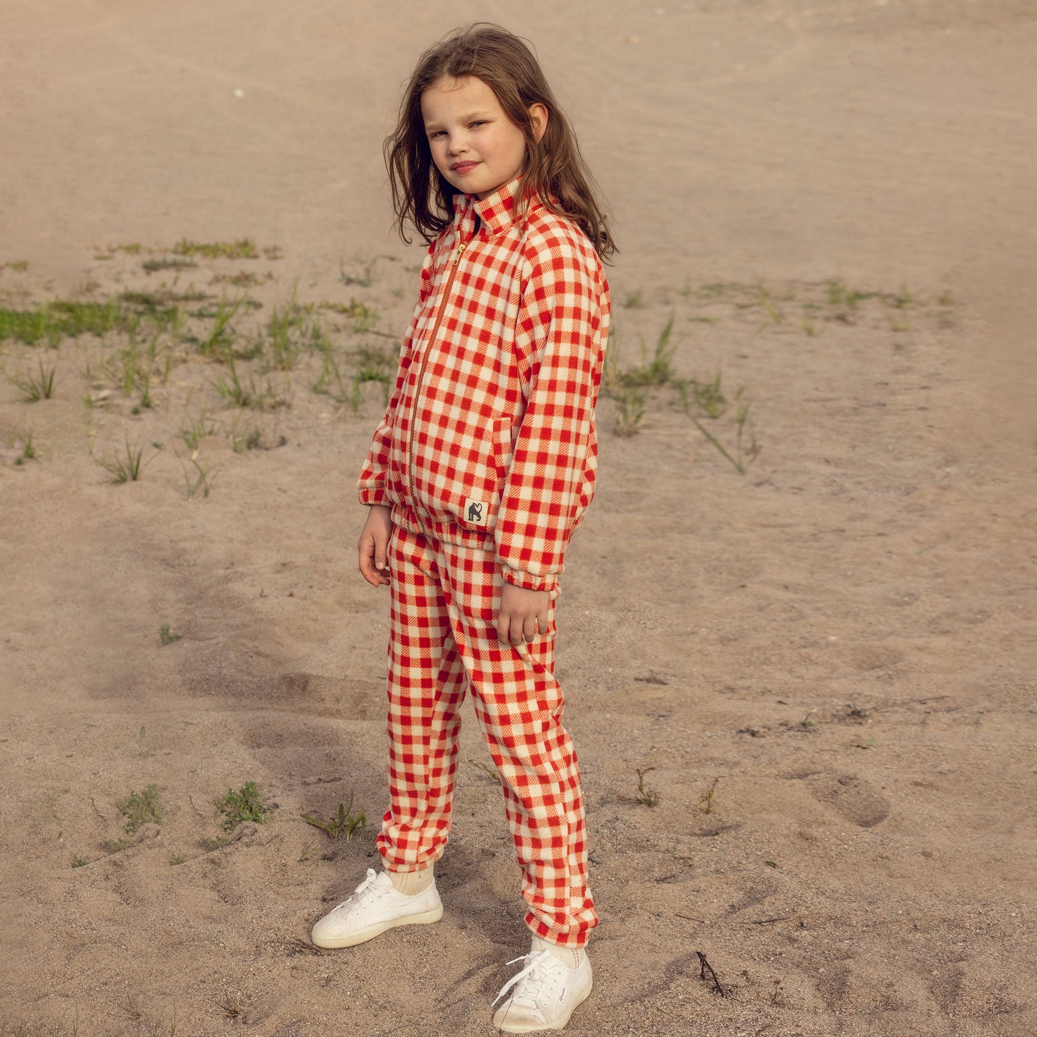 Child wearing a red and white checkered outfit standing on sandy ground. - Little-ish