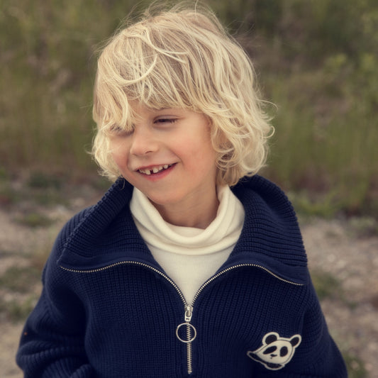Child wearing a navy blue jacket with a logo, standing outdoors with greenery in the background