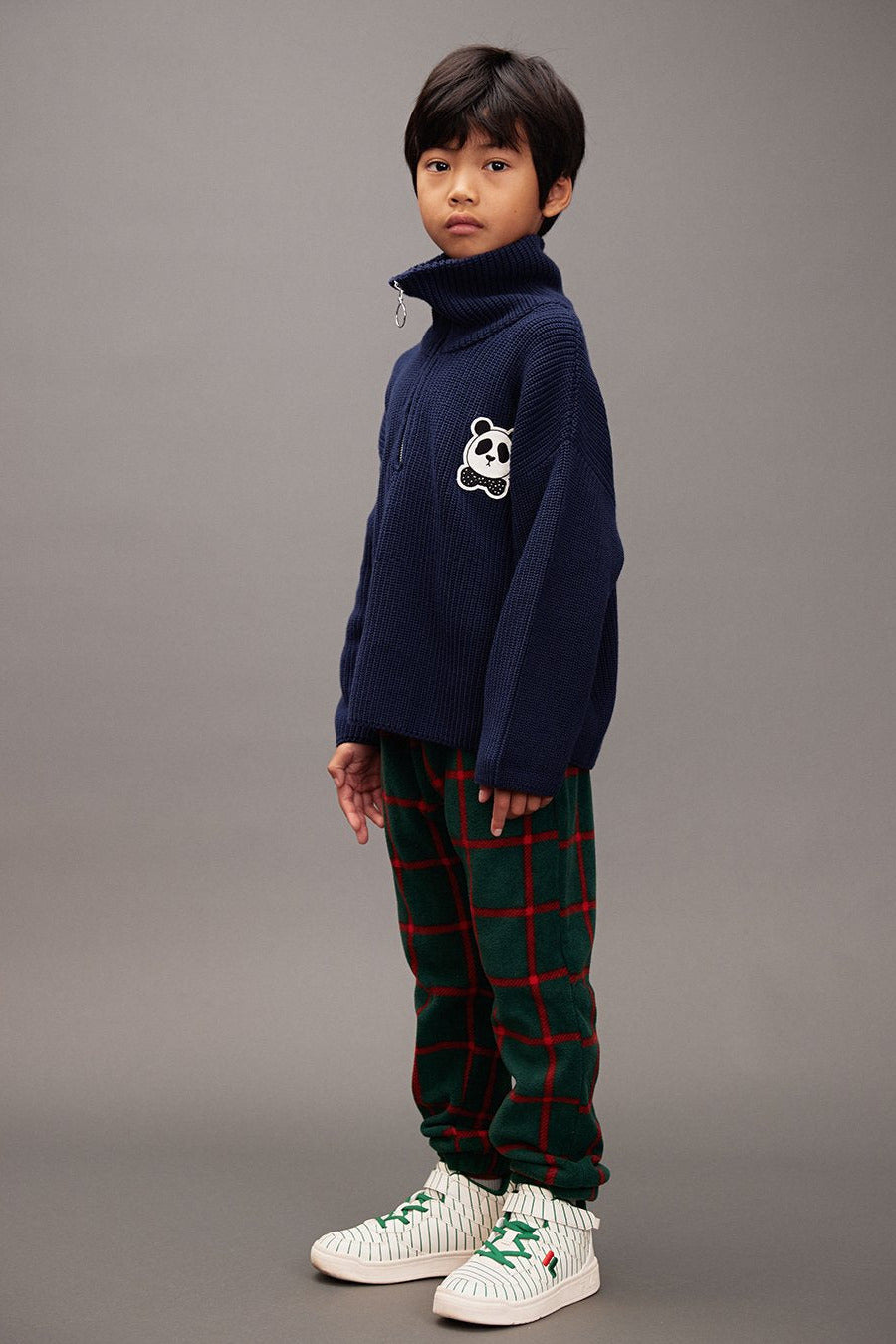 Child wearing a navy sweater with a panda design and plaid fleece pants on a green background - Little-ish