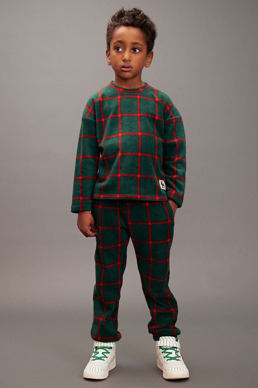 Child wearing a green and red plaid outfit with white sneakers on a gray background - Little-ish