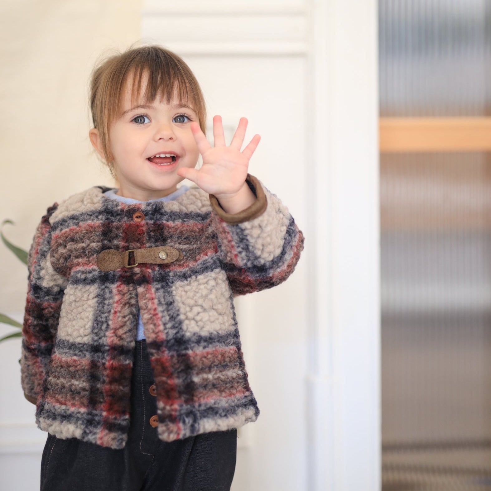 Child wearing a plaid coat waving at the camera indoors. - Little-ish