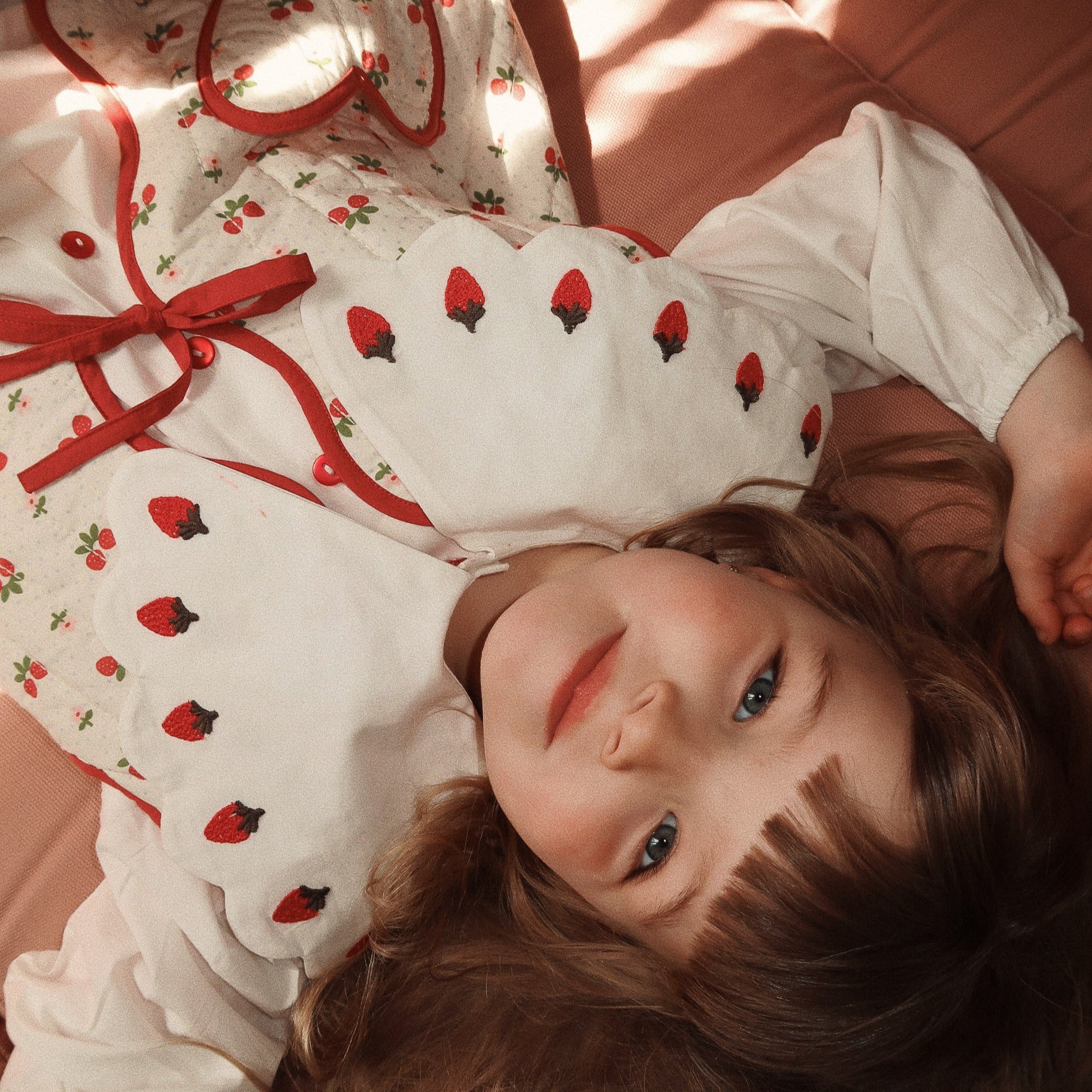 Young girl wearing a white dress with red strawberry pattern lying on a pink surface. - Little-ish
