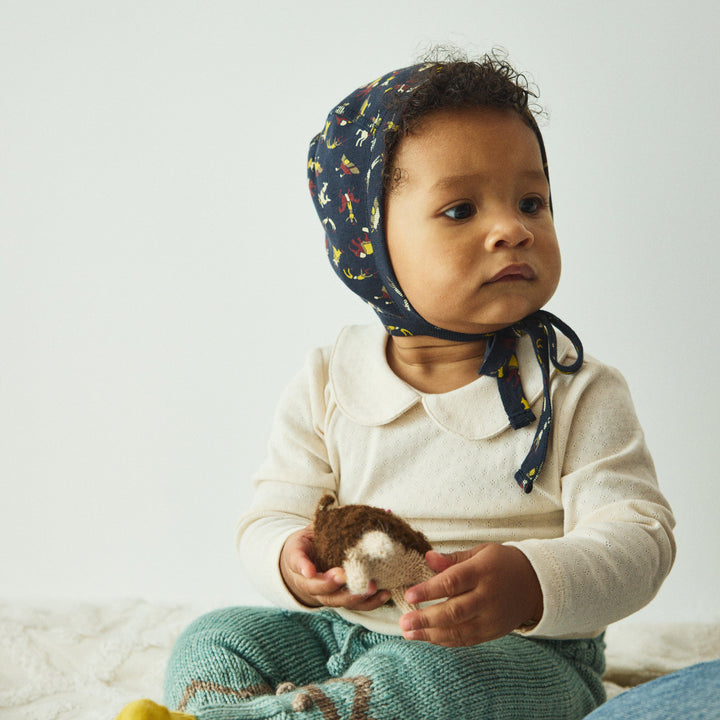 Baby wearing a navy blue bonnet with yellow patterns, holding a small toy, sitting on a white surface.