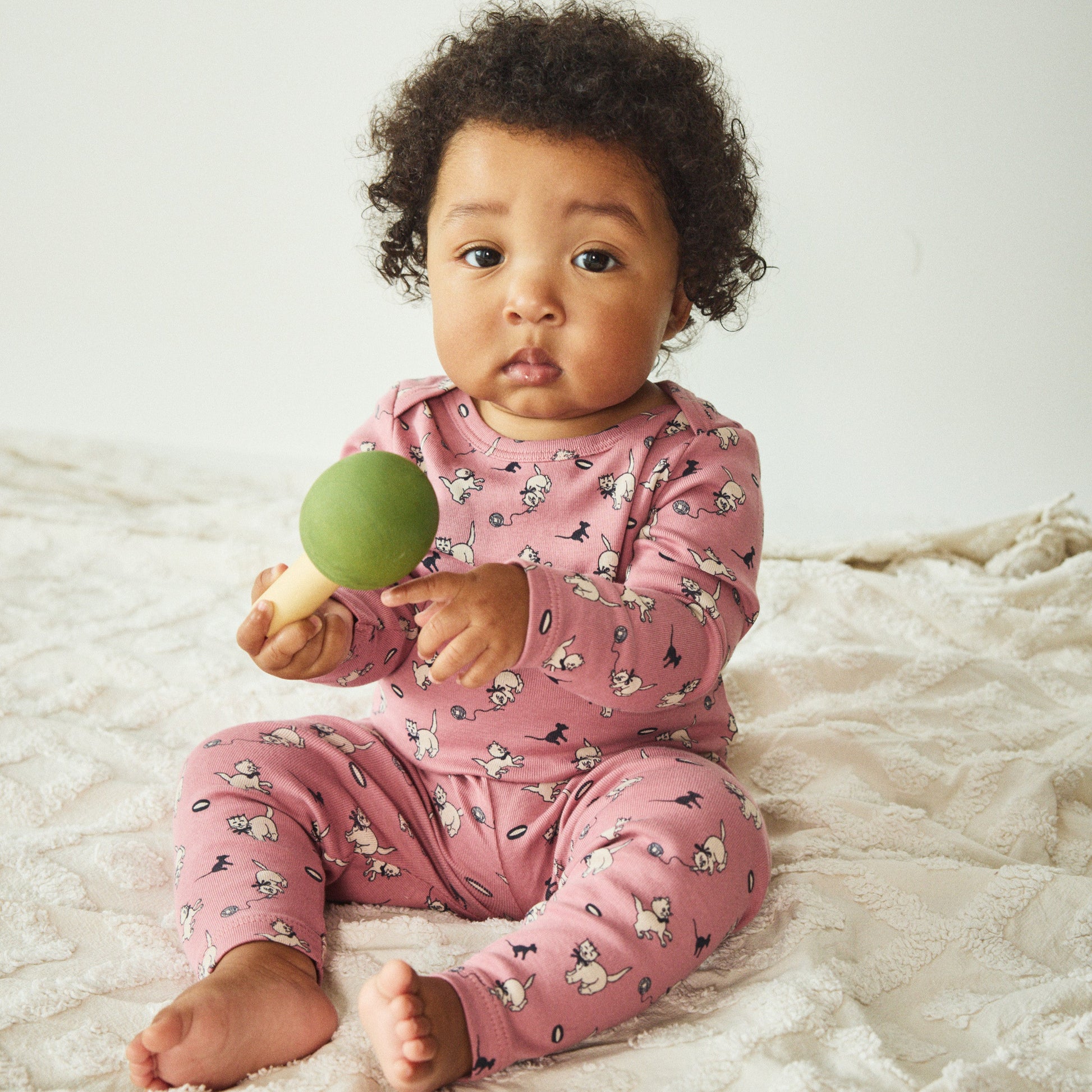 Baby in pink pajamas holding a green ball on a white blanket