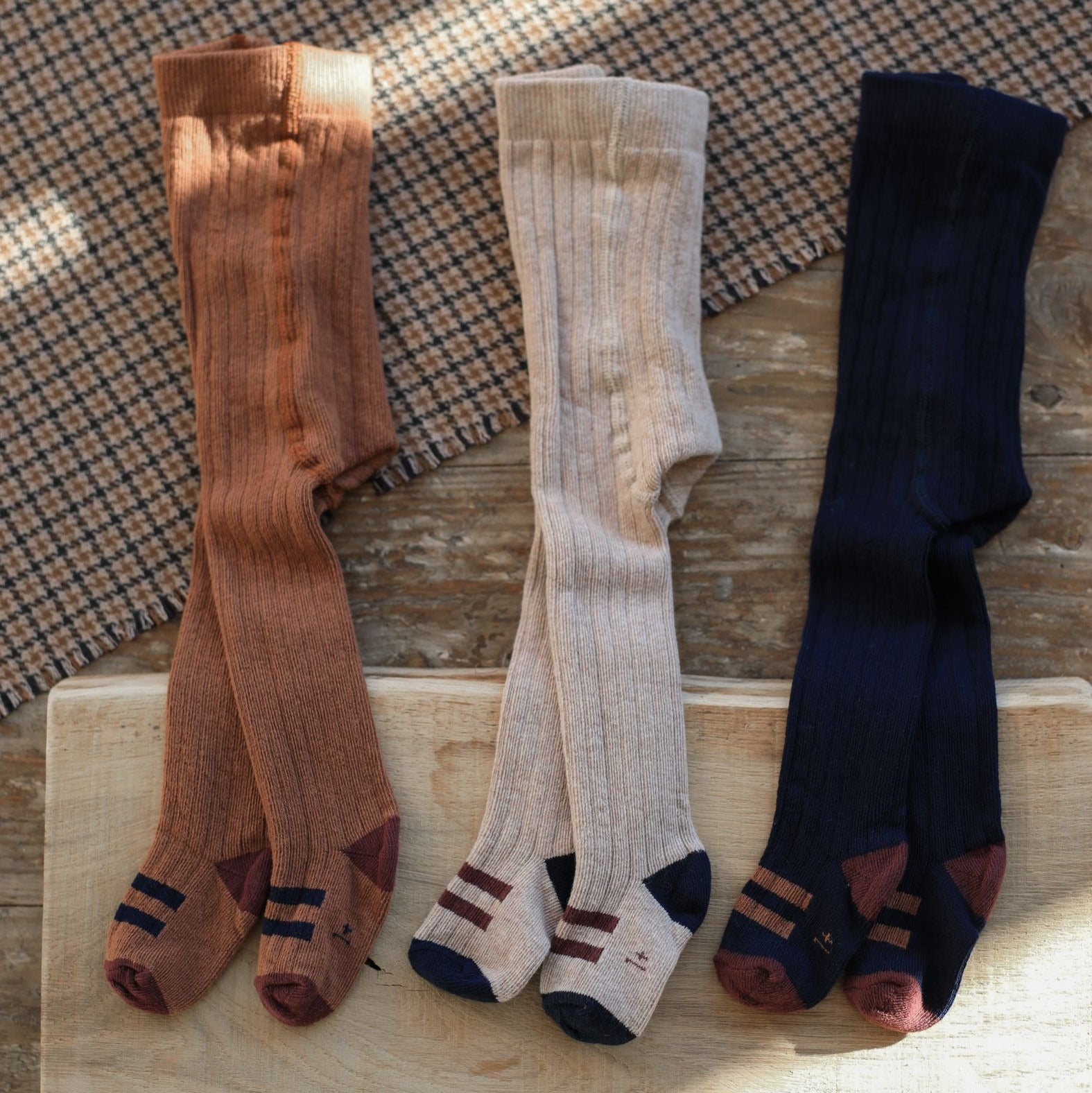 Three pairs of socks in brown, beige, and navy on a wooden surface with a checkered fabric background. - Little-ish