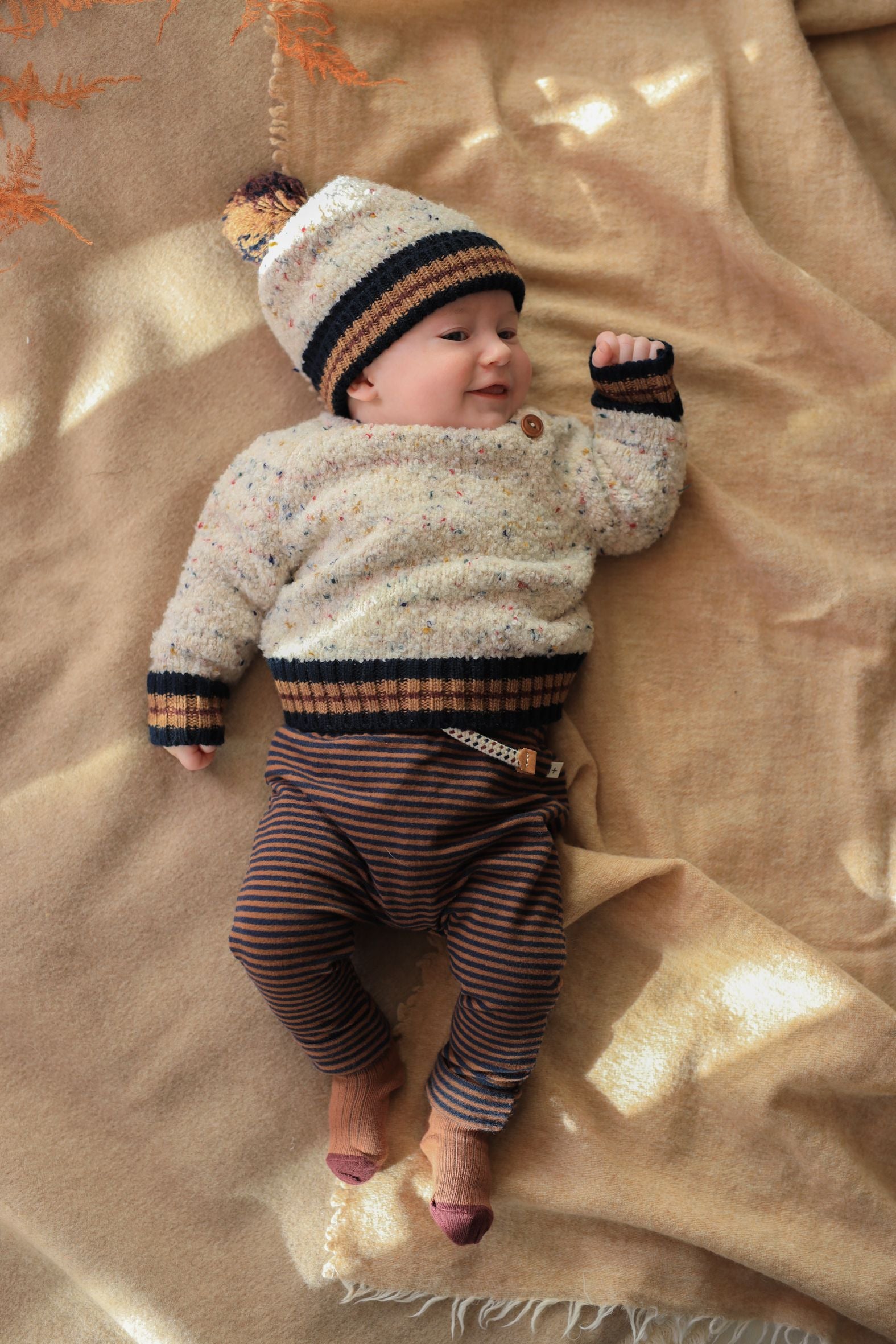 Baby wearing a striped sweater and hat lying on a beige blanket - Little-ish