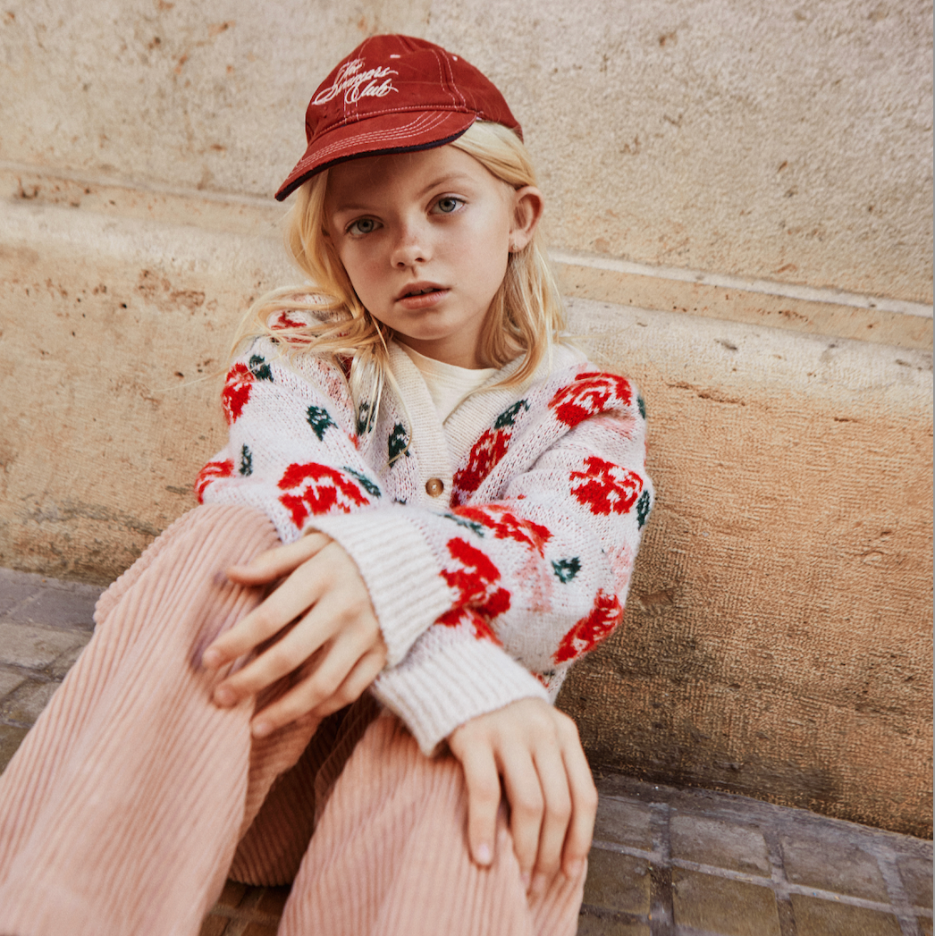 Child wearing a red cap and floral cardigan sitting on a stone bench. - Little-ish