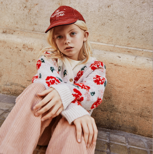 Child wearing a red cap and floral cardigan sitting on a stone bench.