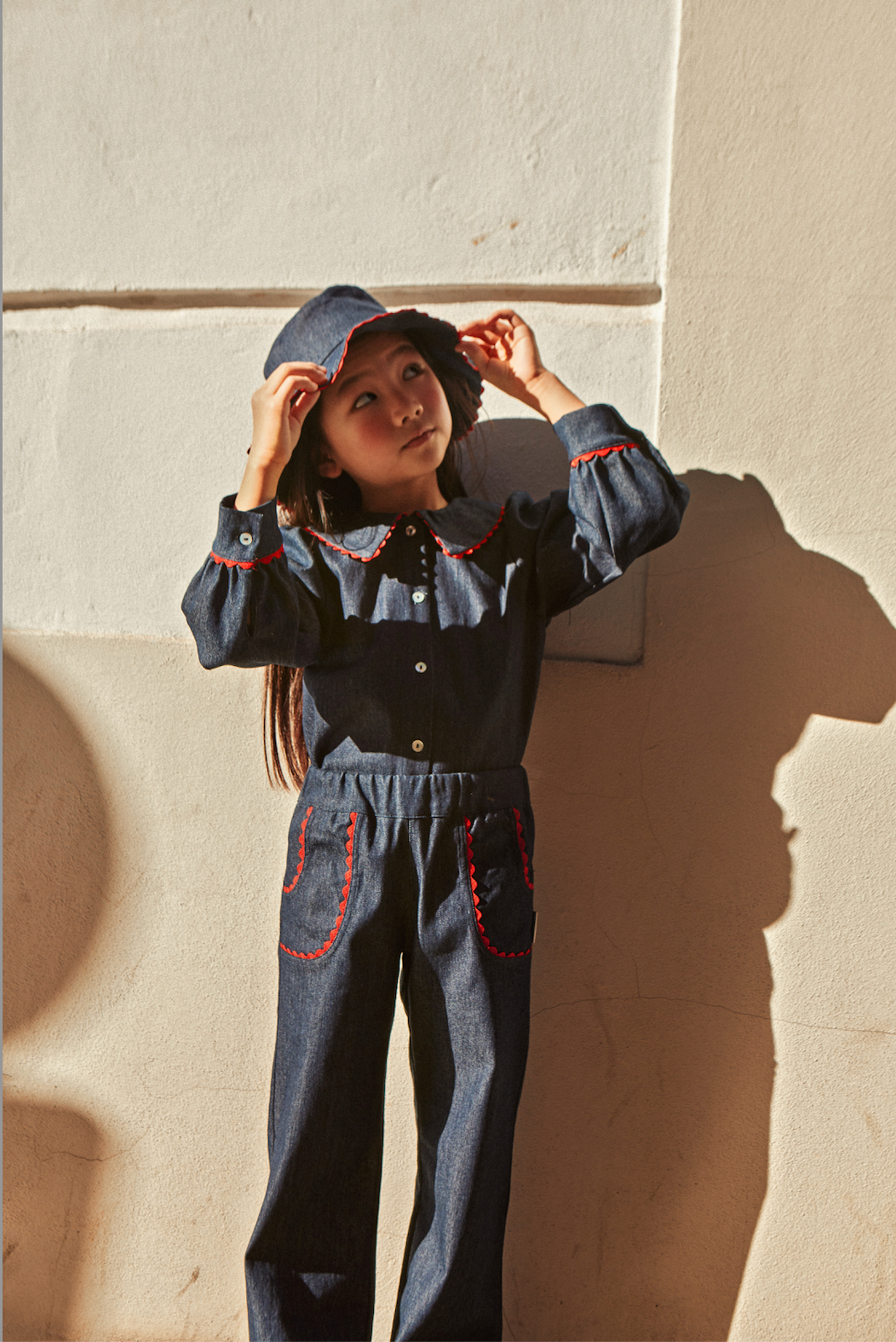 Child wearing a denim outfit with red accents against a light-colored wall.