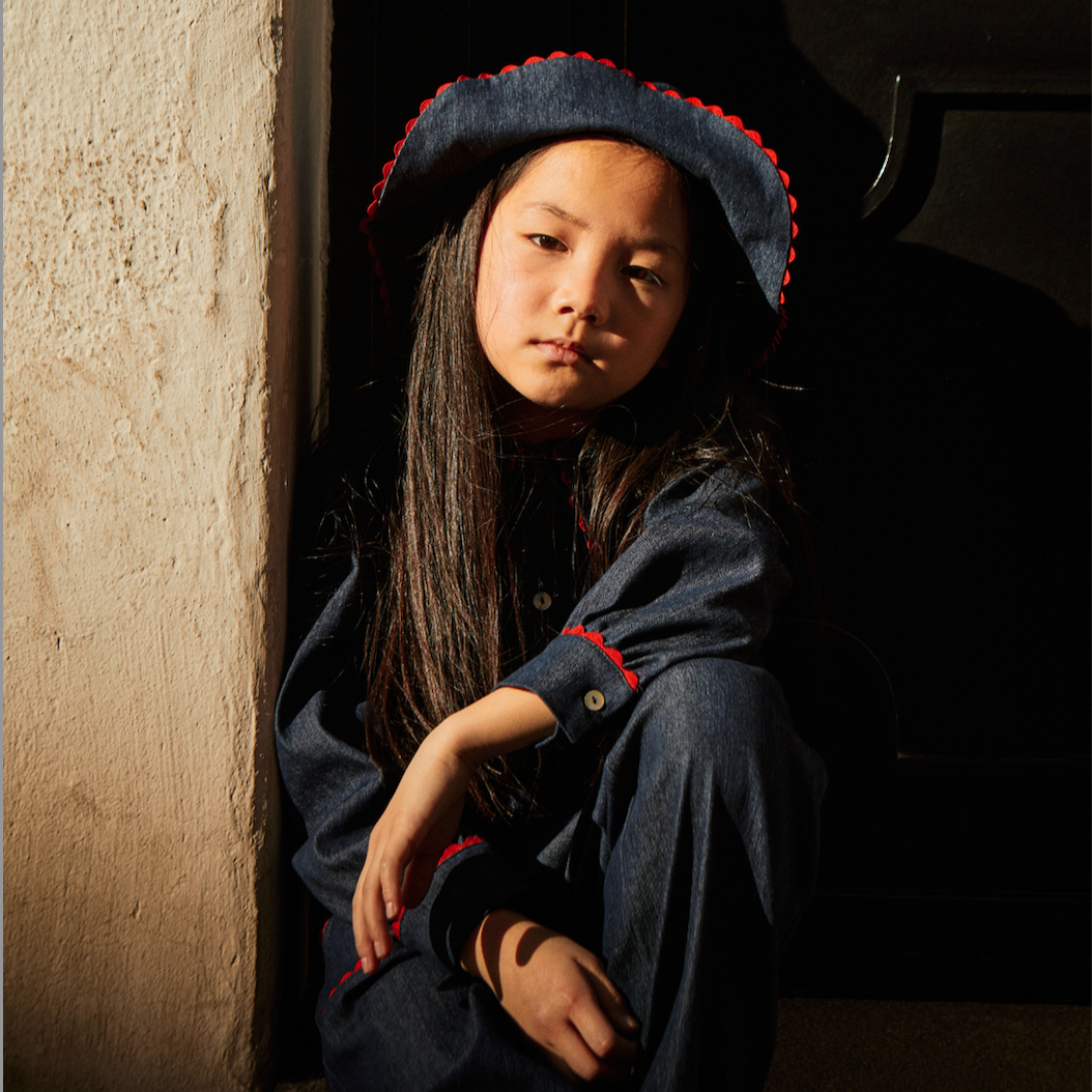 Young girl in a dark outfit with a red trim sitting against a textured wall. - Little-ish