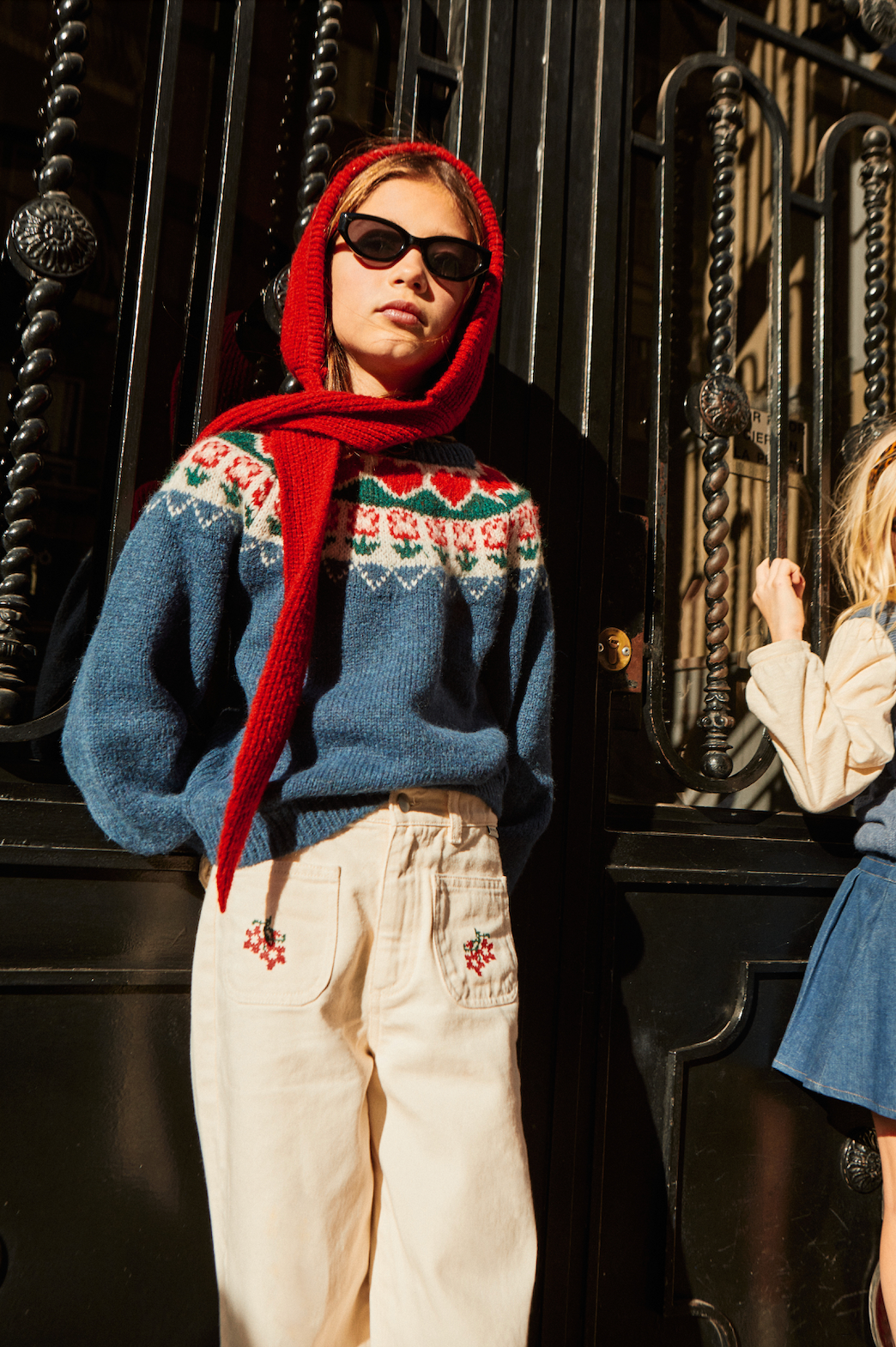 Person wearing a blue sweater with a red scarf and sunglasses, standing in front of a black metal gate. - Little-ish