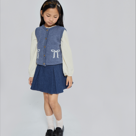 Young girl wearing a denim vest with bow design, skirt, white shirt, and black shoes on a light gray background