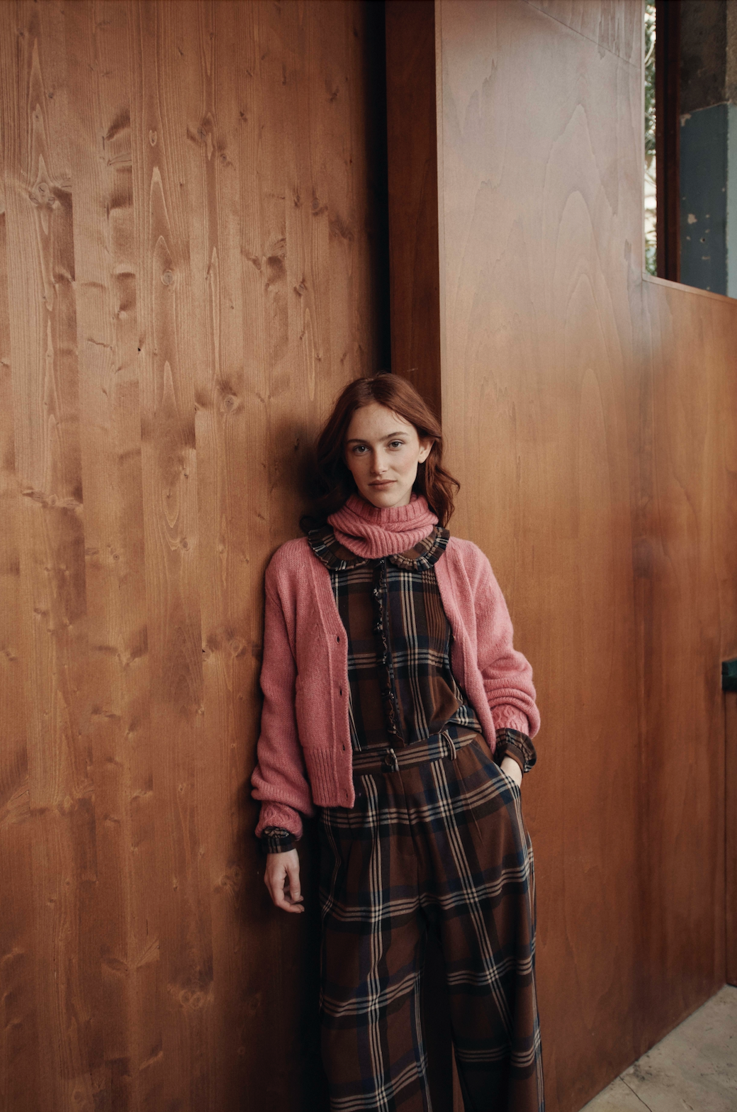 Woman in a pink cardigan and plaid dress standing against a wooden wall. - Little-ish