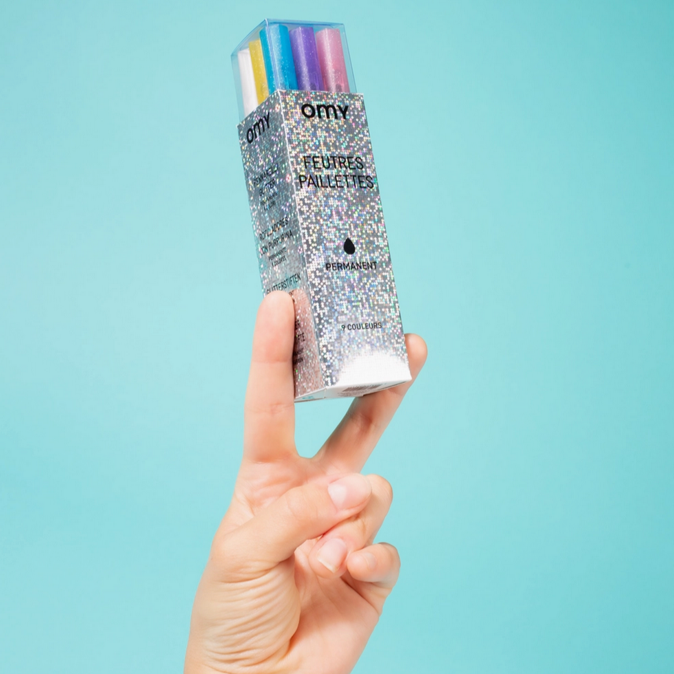 Hand holding a glittery box of OMY Feathers against a light blue background