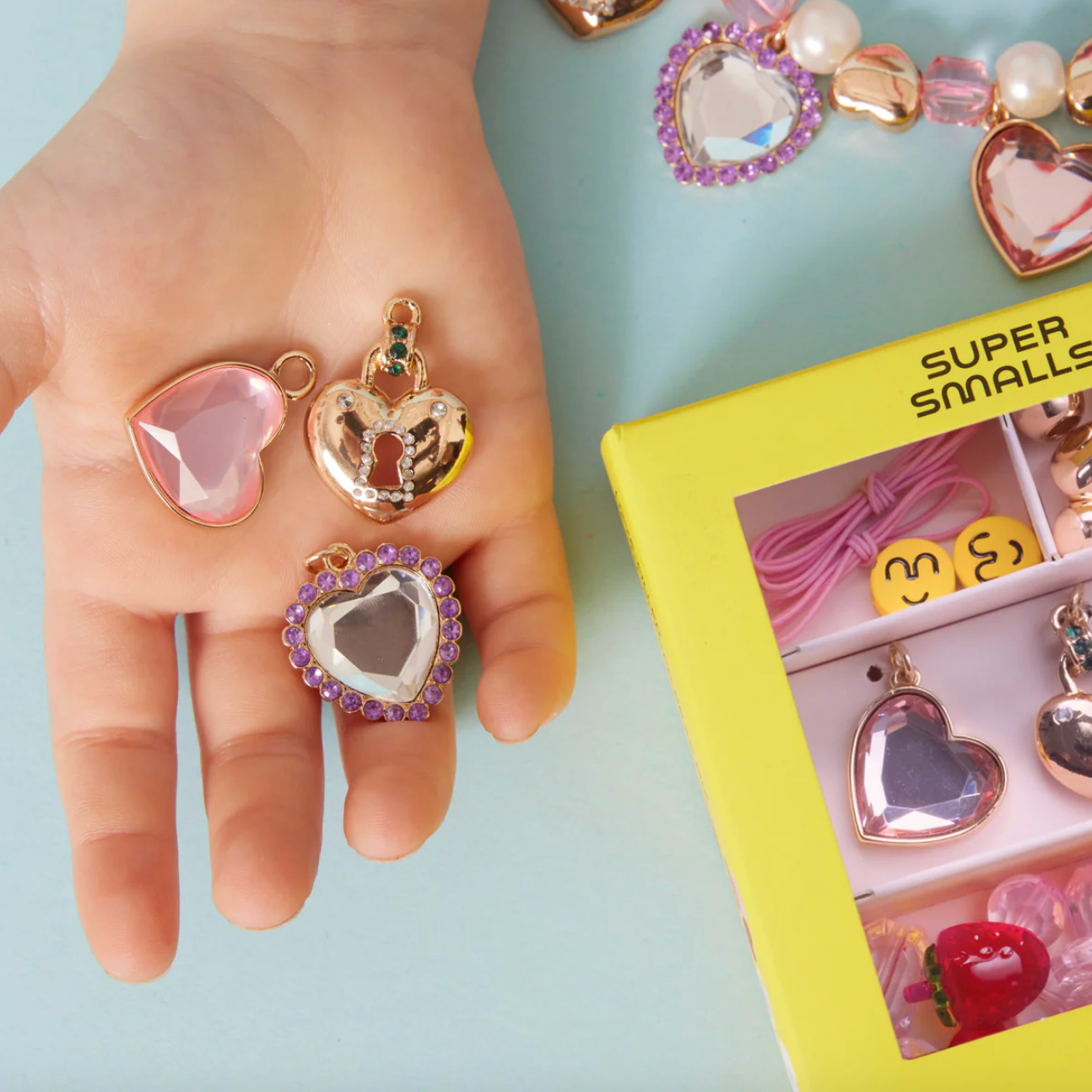 Heart-shaped jewelry charms on a hand with a box labeled 'Super Smalls' in the background.