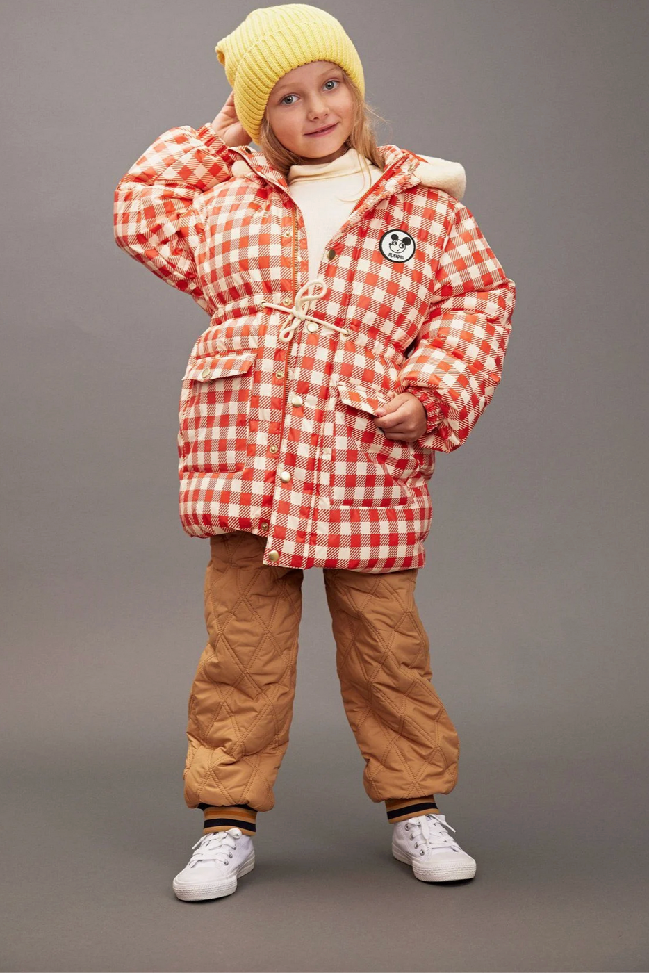 Child wearing a red and white checkered coat with a yellow beanie and brown pants on a gray background - Little-ish
