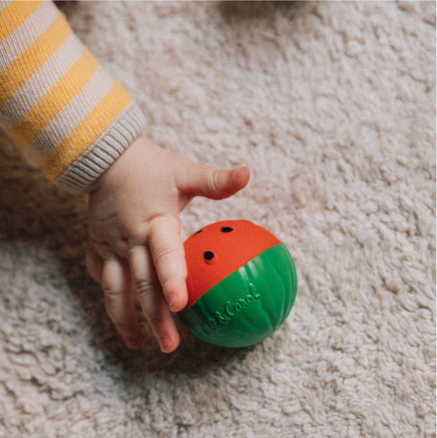 Child's hand holding a colorful ball with 'Bob & Co.' branding on a carpeted floor. - Little-ish