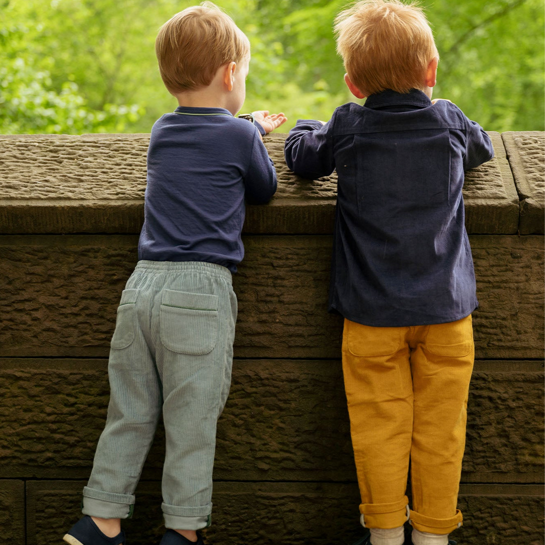 Two children standing on a wooden platform looking over a railing with greenery in the background. - Little-ish