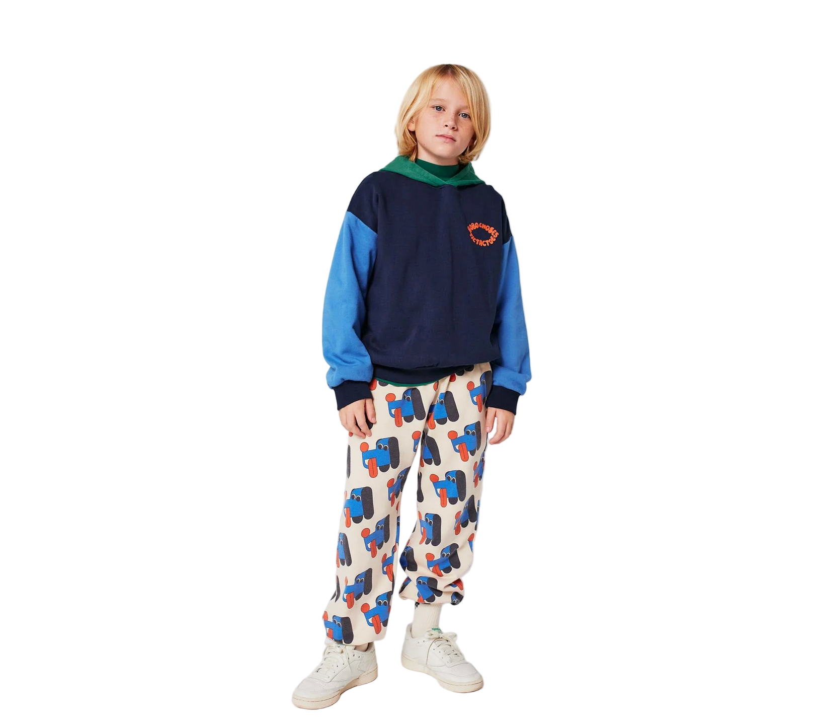 Child wearing a blue and black sweater with a colorful patterned shirt underneath, white sneakers, and multicolored pants on a plain background. - Little-ish