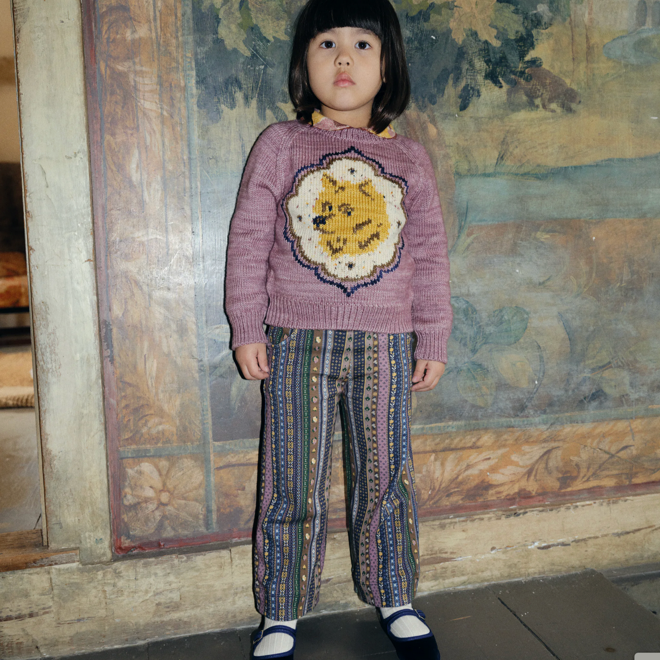 Child wearing a pink sweater with a colorful design and patterned pants standing in front of a textured wall.