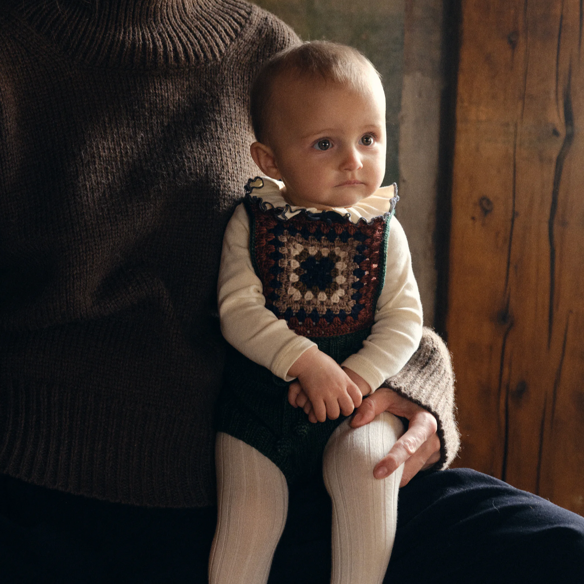 Baby wearing a crocheted outfit sitting on an adult's lap with a wooden background