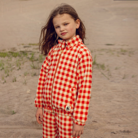 Young girl wearing a red and white checkered outfit standing on sandy ground.