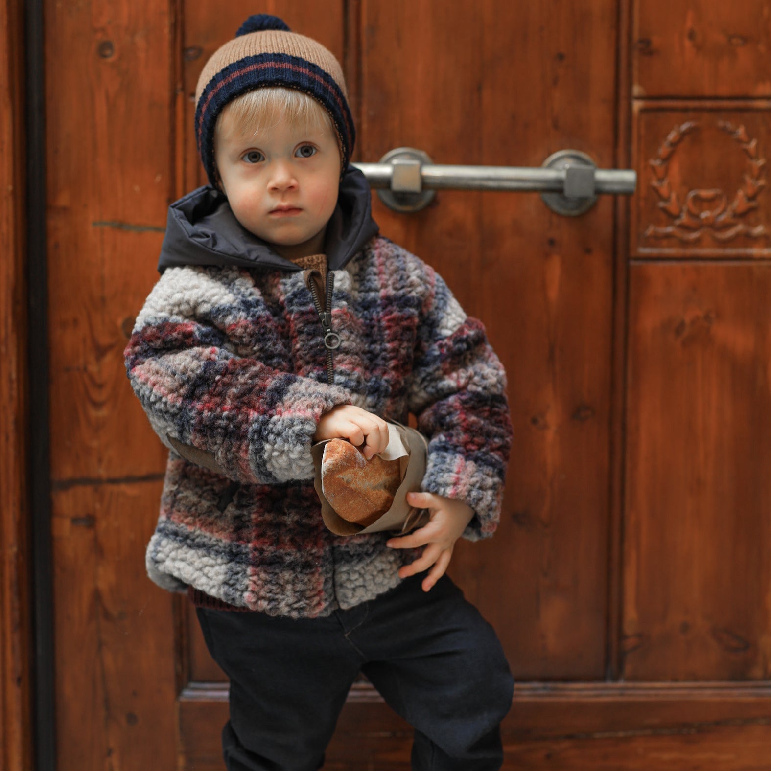 Child wearing a patterned coat and hat standing in front of a wooden door. - Little-ish