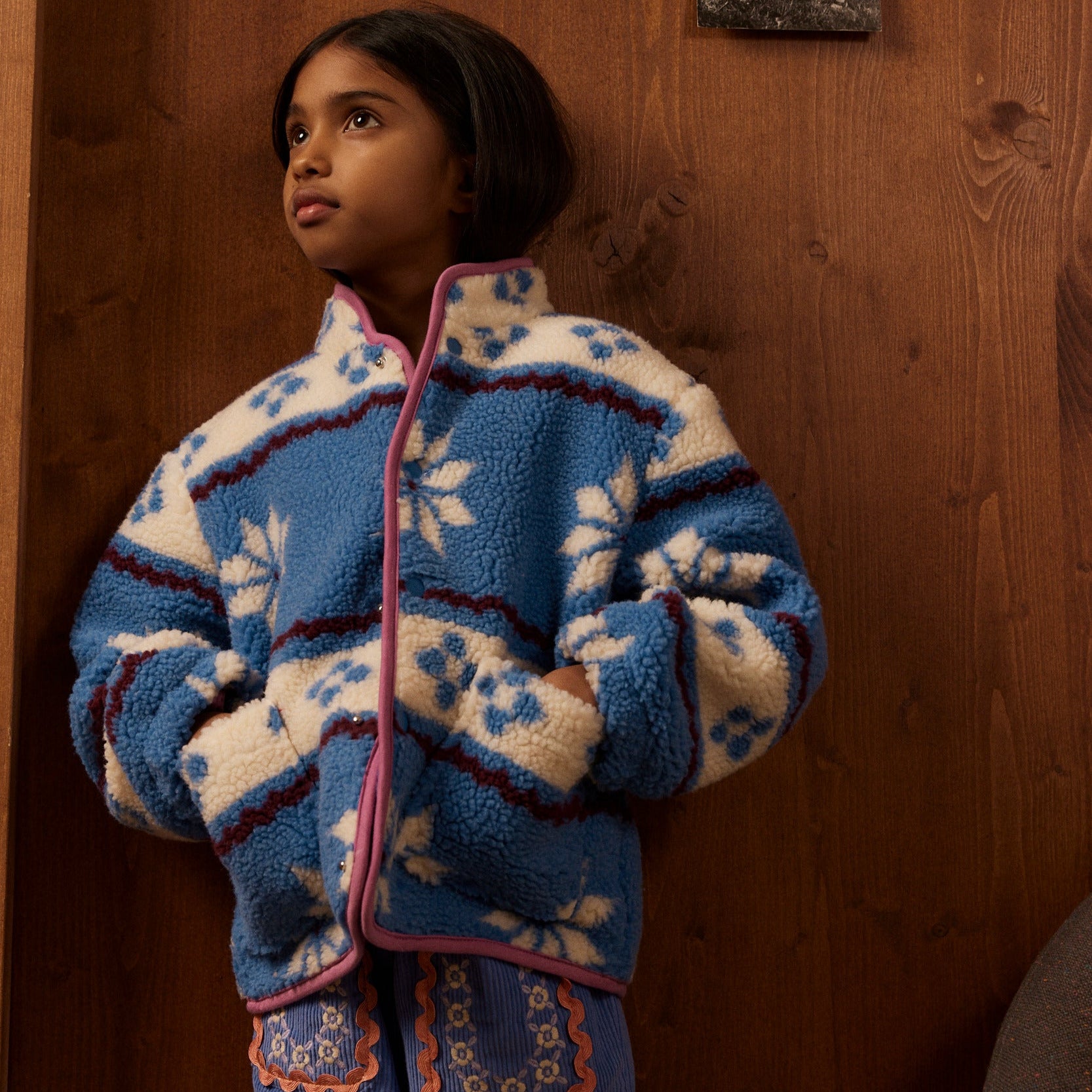 Child wearing a blue and white patterned sweater standing in front of a wooden wall with posters.