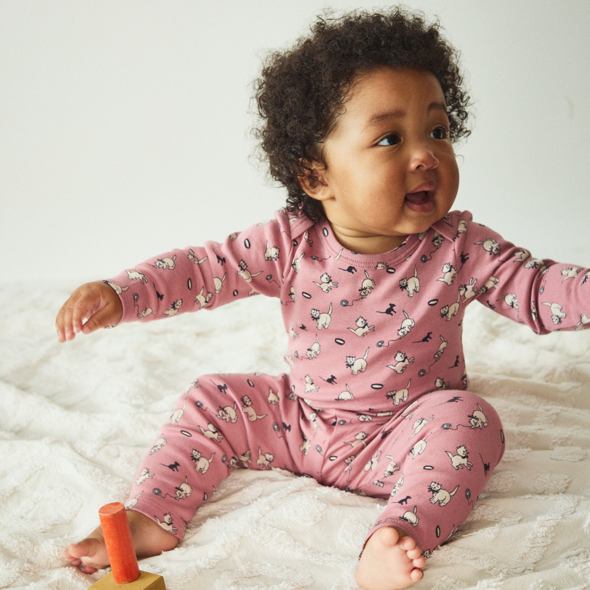 Baby in pink pajamas sitting on a white blanket with a wooden toy.