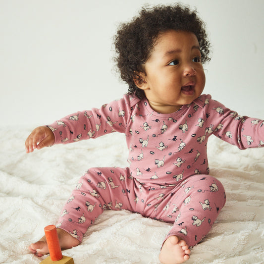 Baby in pink pajamas sitting on a white blanket with a wooden toy.