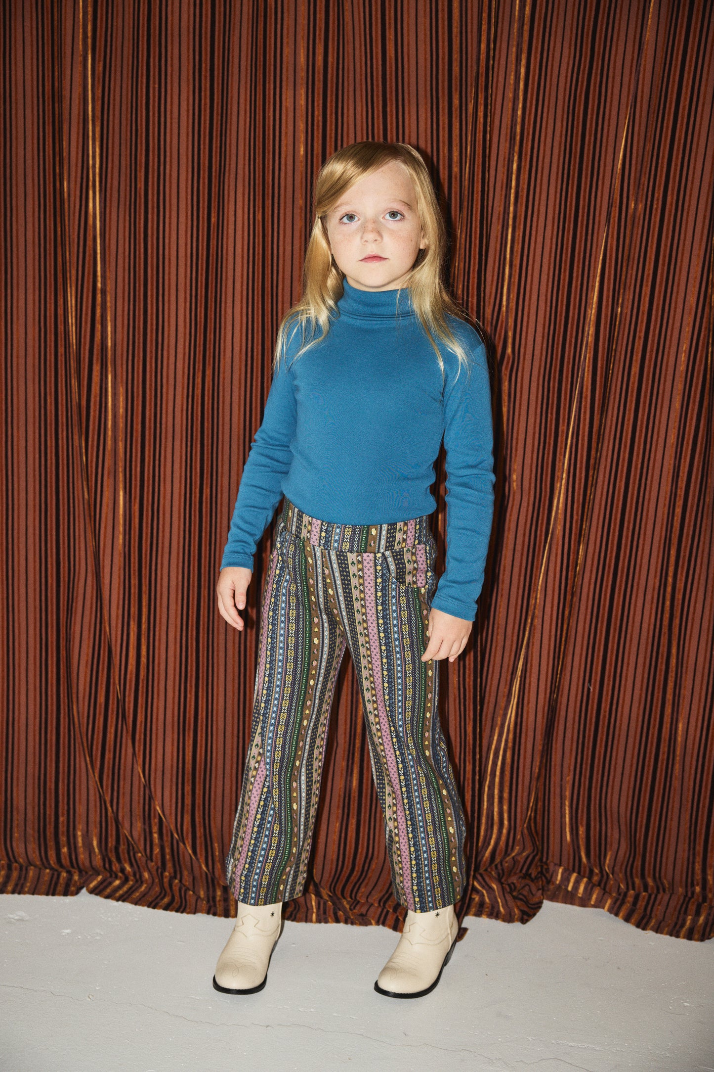 Child wearing a blue turtleneck and patterned pants standing against a brown curtain.
