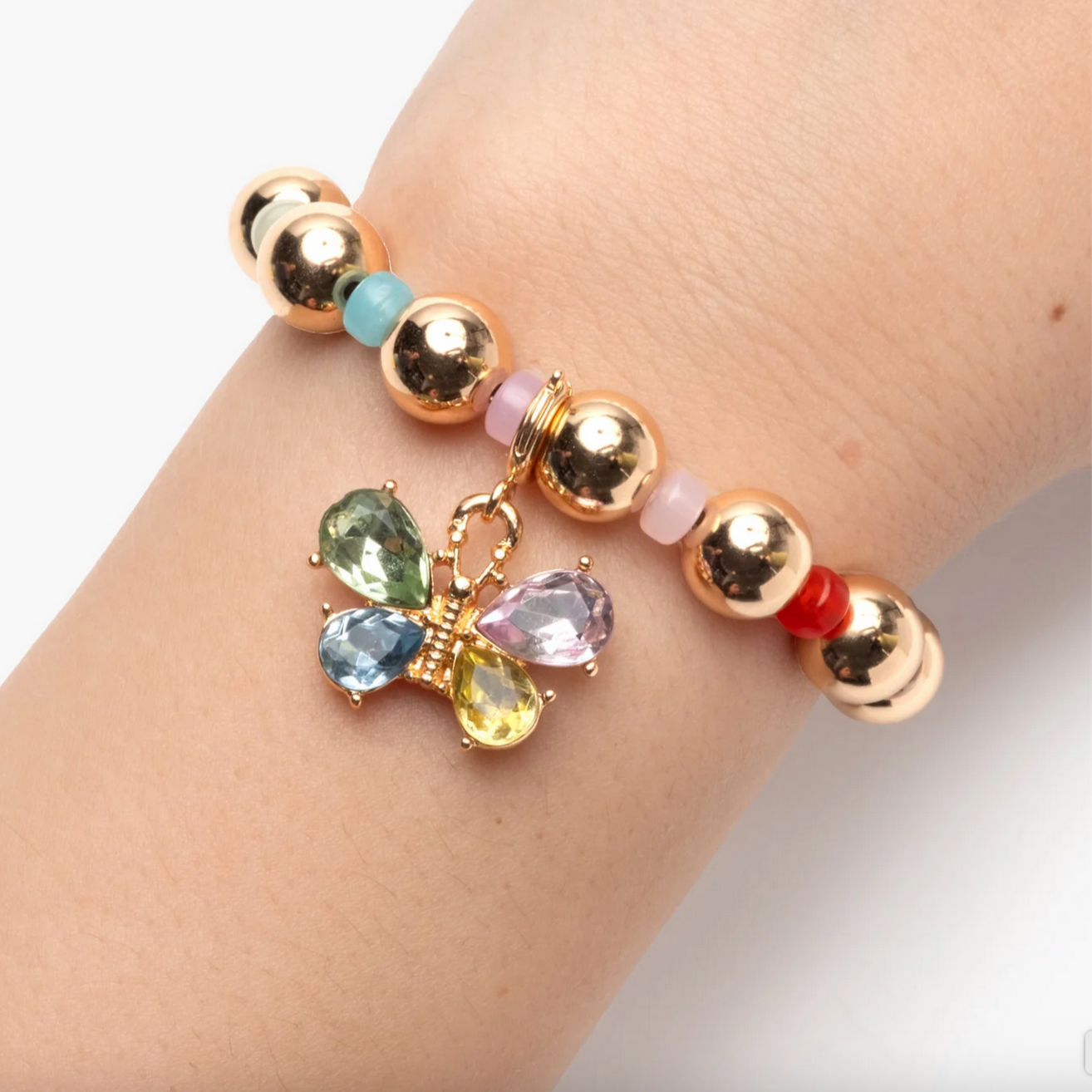 Gold beaded bracelet with a colorful butterfly charm on a wrist against a white background - Little-ish