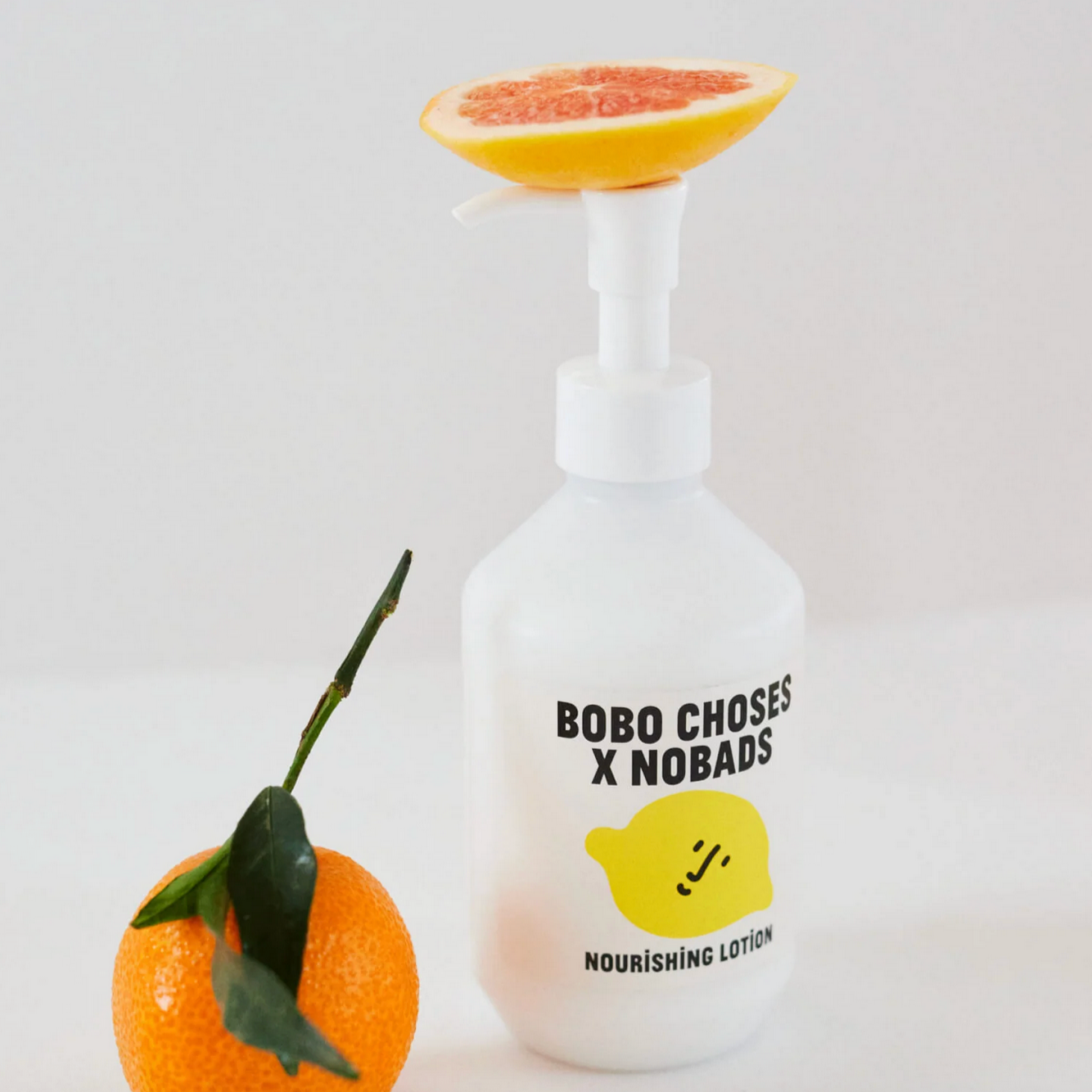 White lotion bottle with 'Bobo Choses x Nobads' branding next to a grapefruit on a light gray background - Little-ish