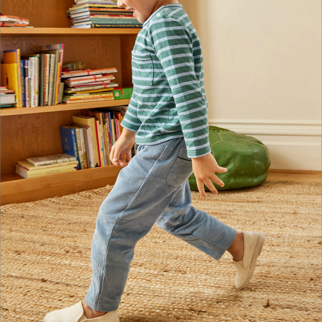 Child walking in a room with a bookshelf and green bag in the background - Little-ish