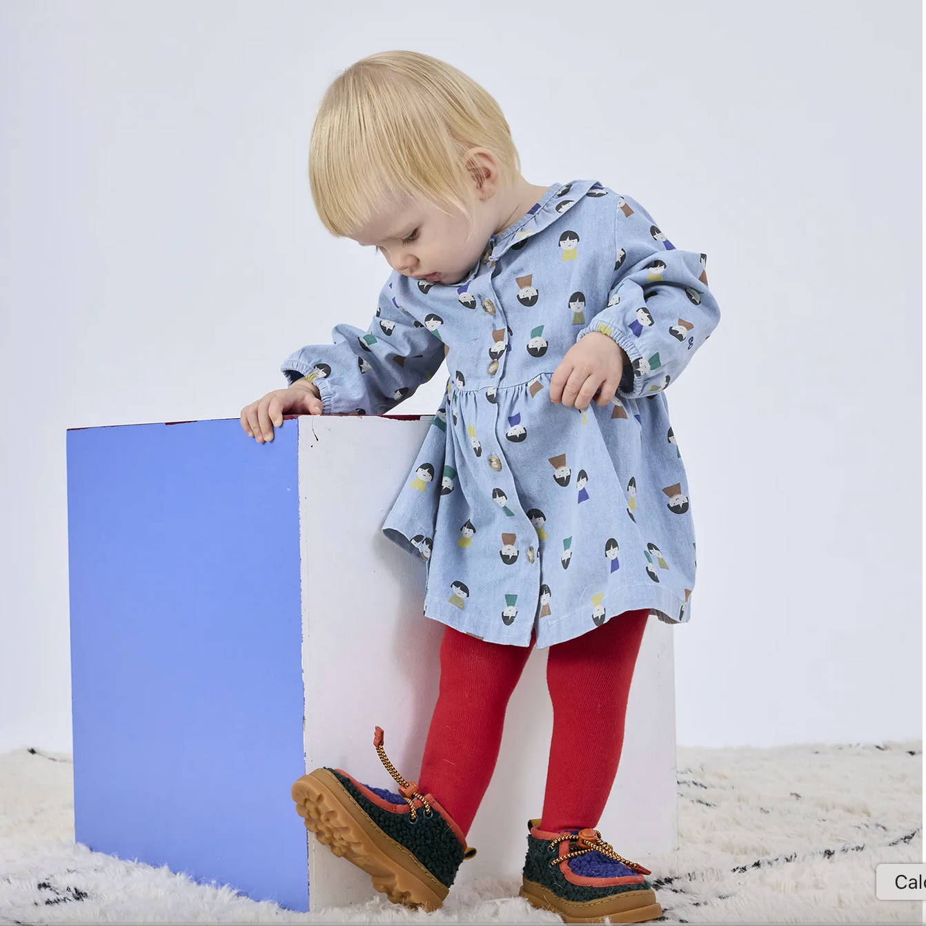 Child wearing a patterned raincoat and red leggings standing next to a blue and white block on a white background.