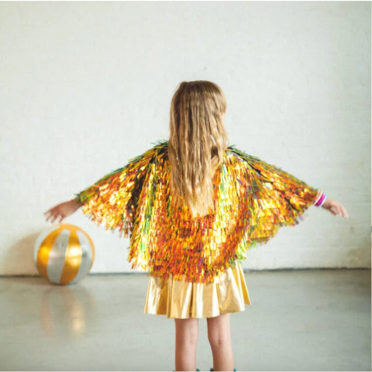 Child wearing a colorful sequin dress with arms outstretched, standing against a plain background.