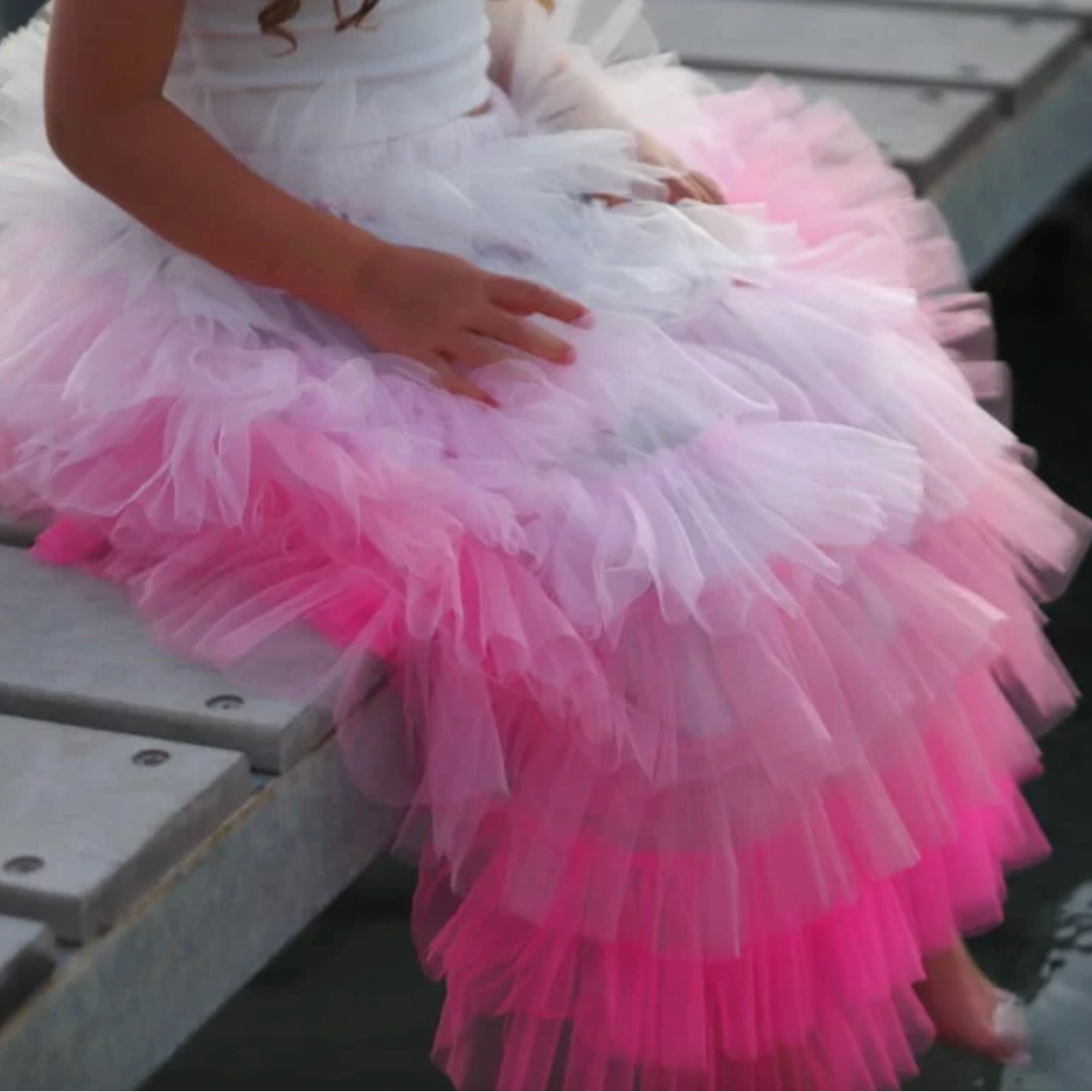 Person wearing a white and pink layered dress sitting on a wooden bench. - Little-ish