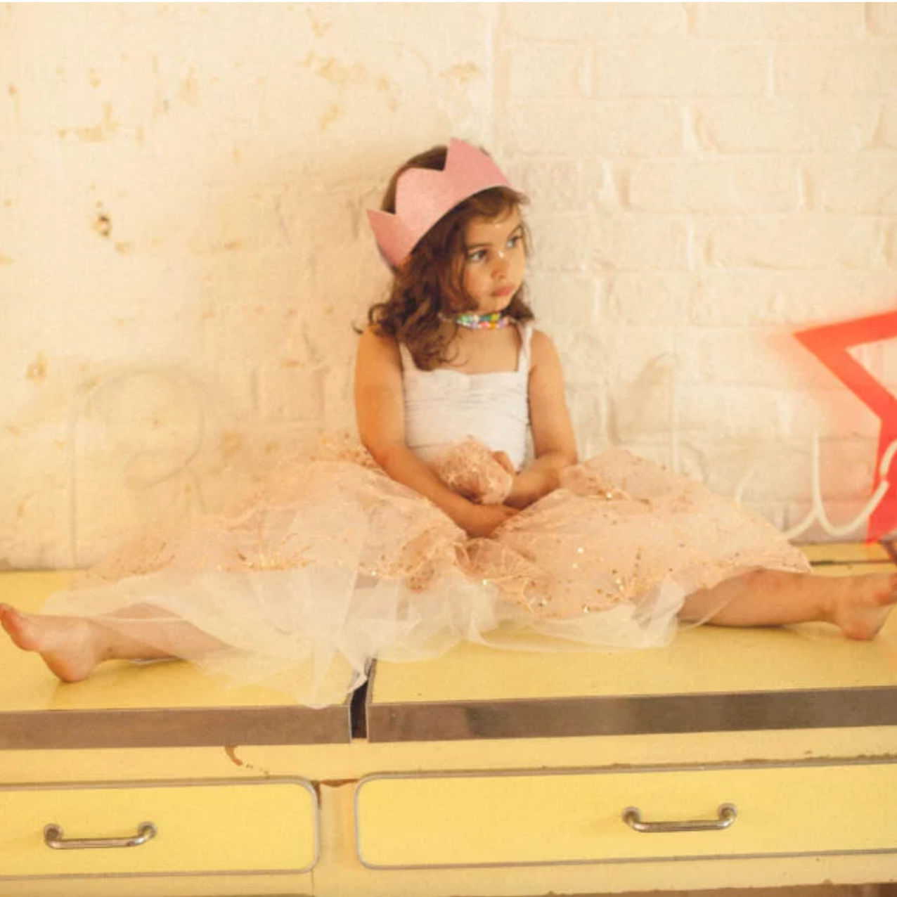 Young girl in a white dress with pink floral details sitting on a yellow vintage suitcase.
