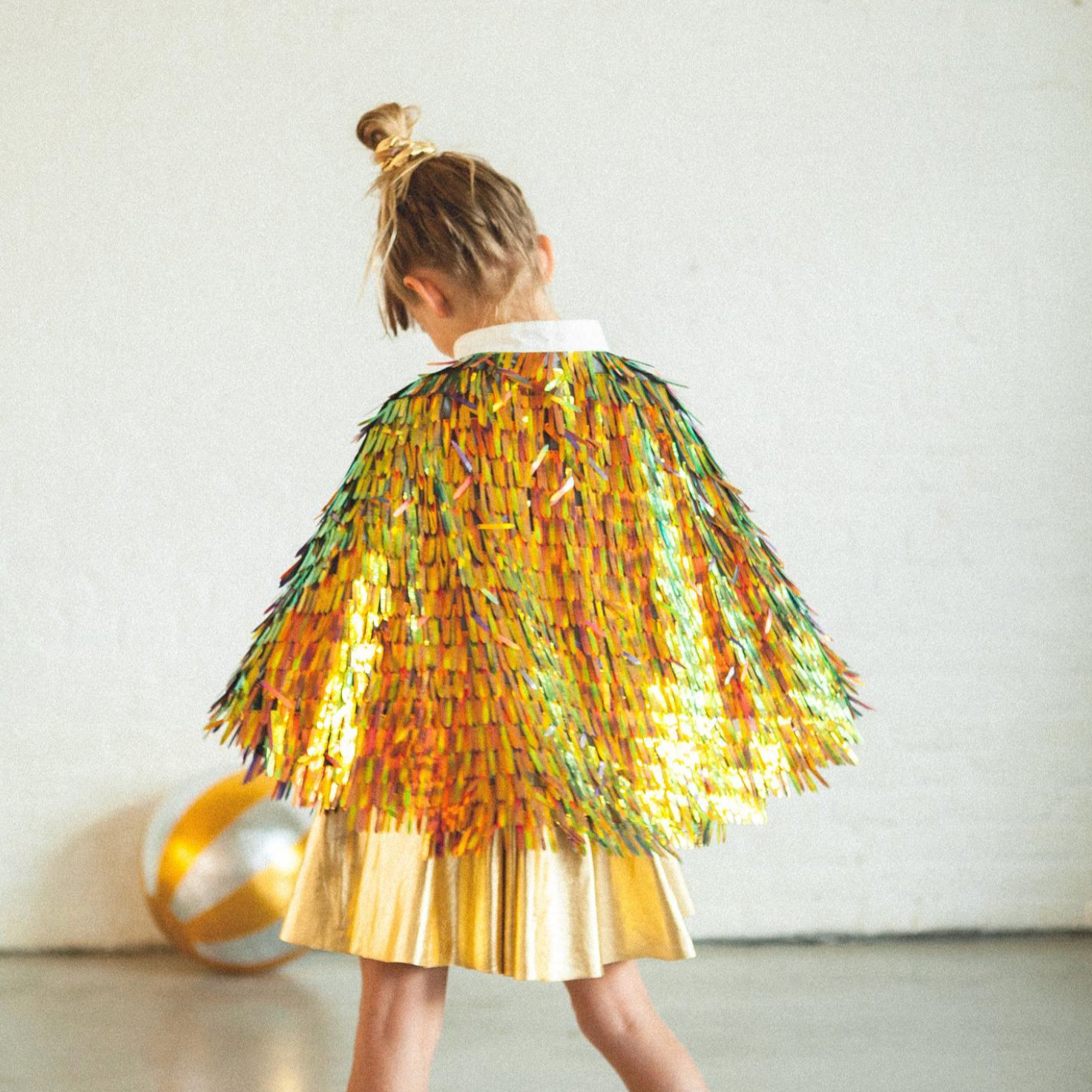 Child wearing a colorful sequin cape and gold skirt against a white background - Little-ish