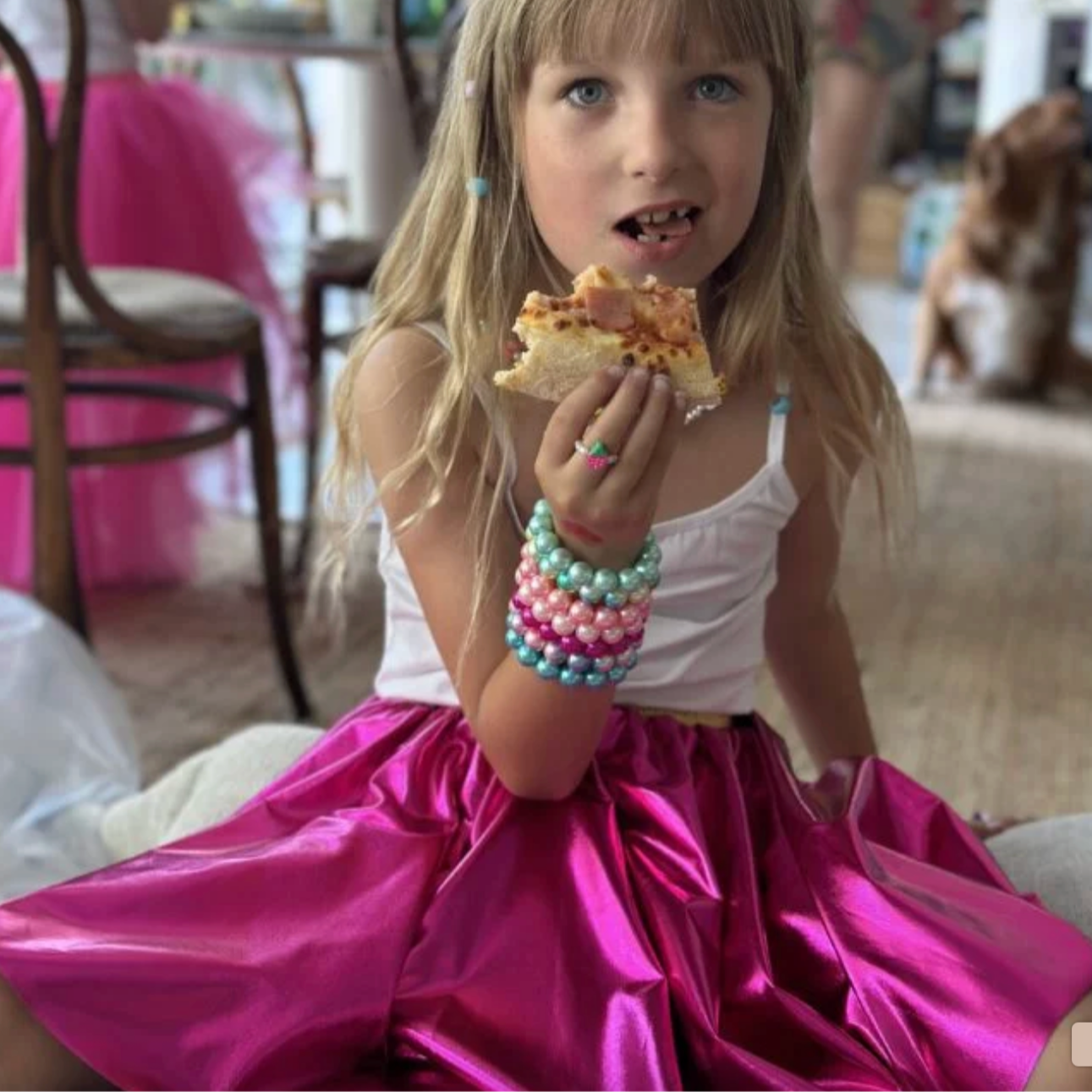 Young girl in a pink skirt and white top holding a piece of pizza indoors. - Little-ish