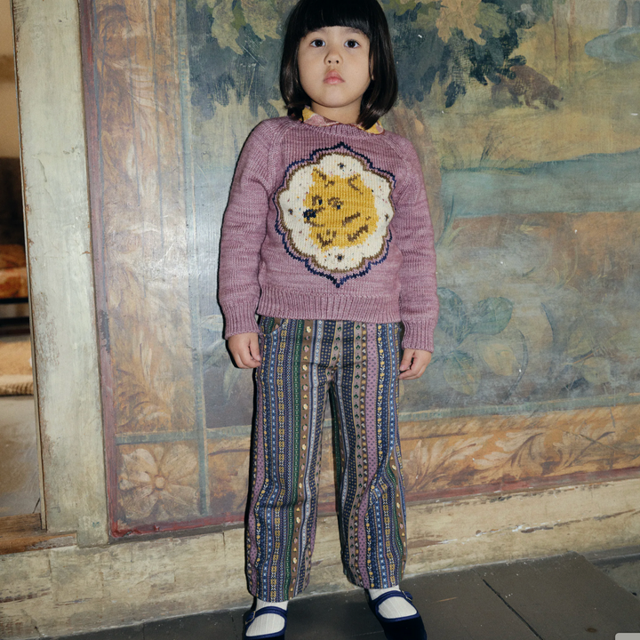 Child wearing a pink sweater with a colorful design and patterned pants standing in front of a textured wall.