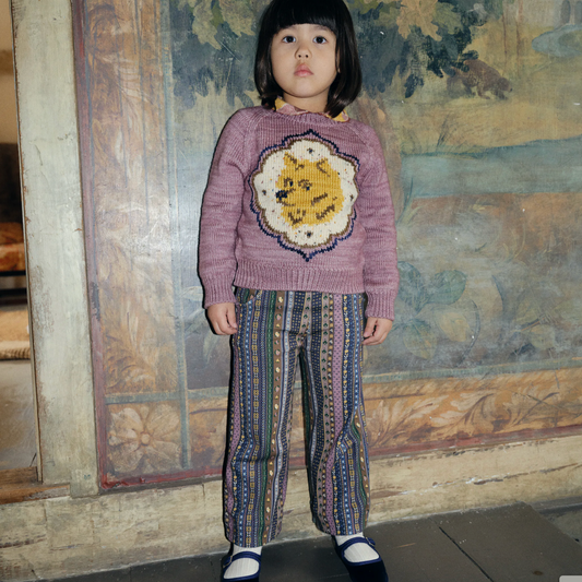Child wearing a pink sweater with a colorful design and patterned pants standing in front of a textured wall.