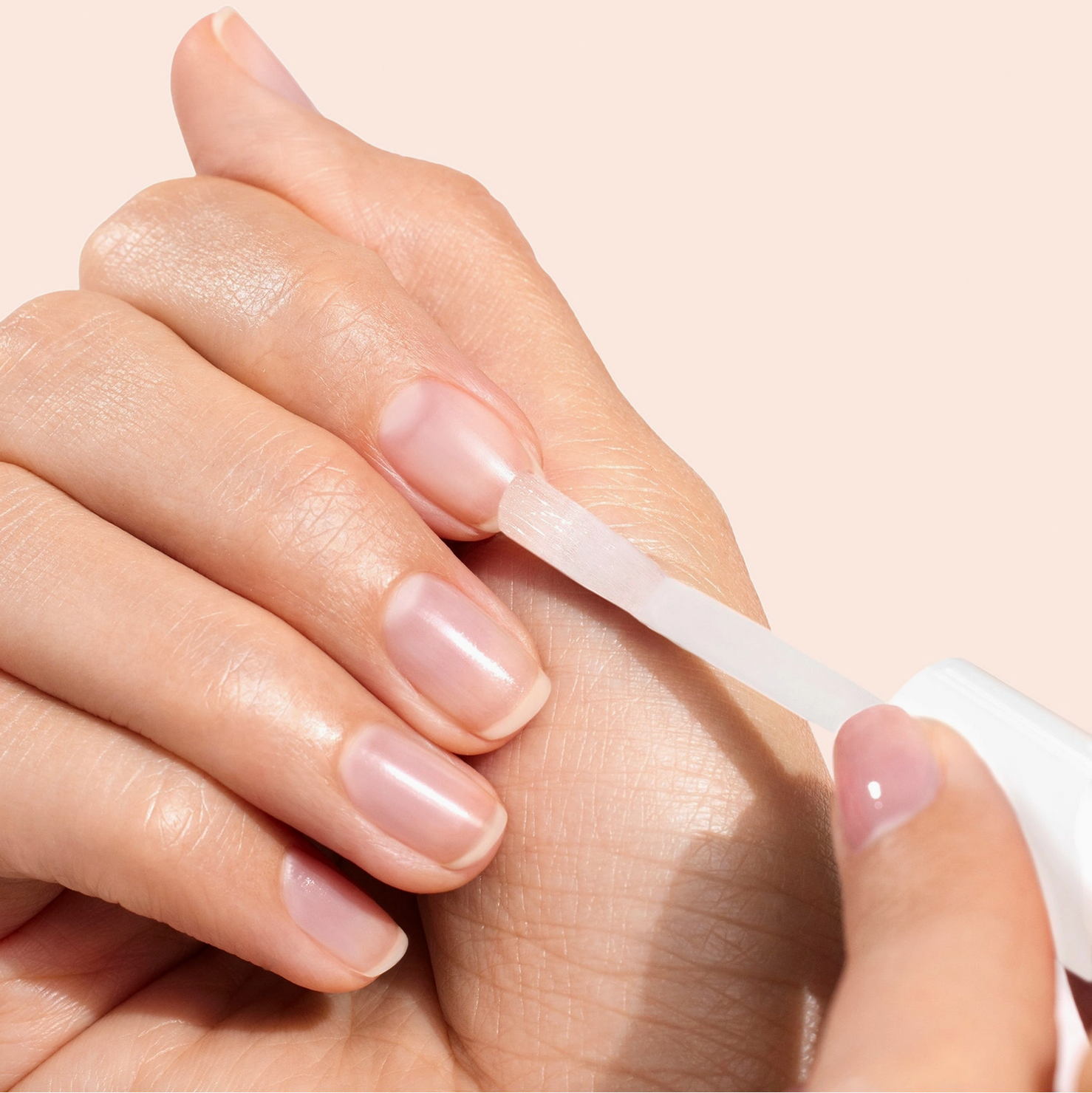Close-up of a hand applying a clear nail polish strip to another hand with a neutral background - Little-ish