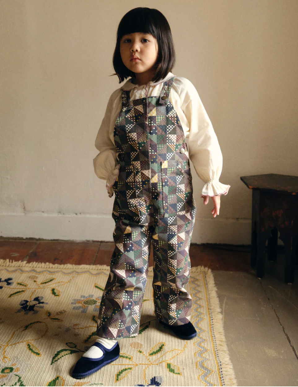 Child wearing a patterned outfit standing on a rug with a neutral background - Little-ish