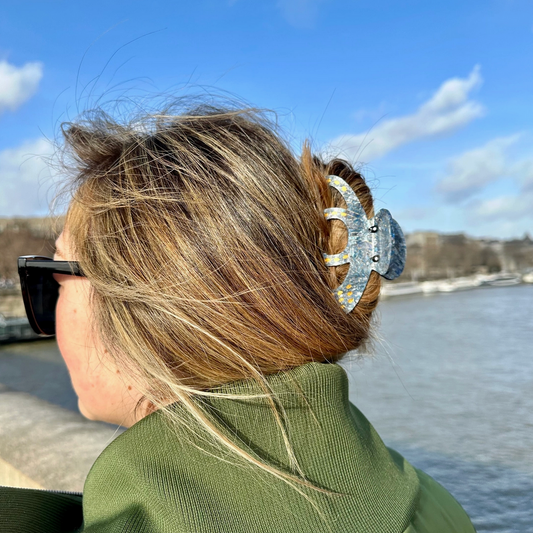 Person with styled hair and sunglasses, standing by a body of water with a blue sky.