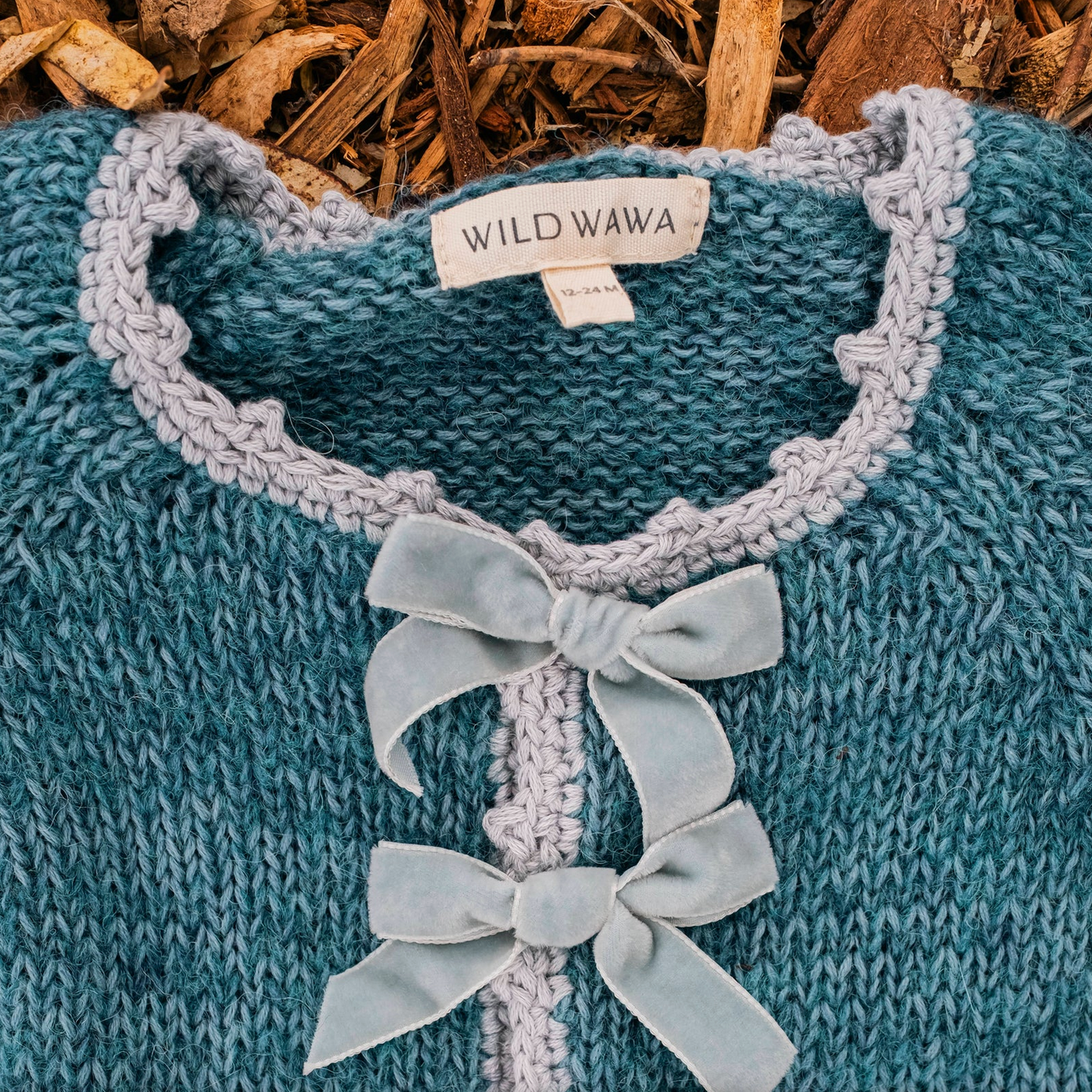 Blue knitted sweater with a collar and bows, featuring 'WILD WAWA' label, on a wooden background.