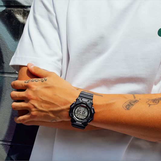 Person wearing a digital watch with a white shirt and tattooed arm against a graffiti-covered wall.