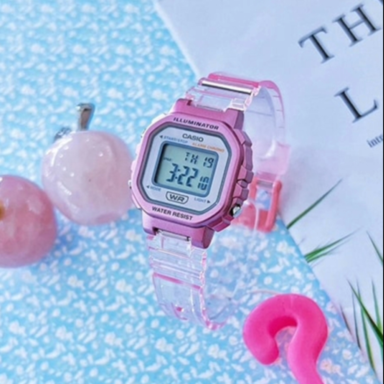 Pink Casio digital watch on a light blue textured background with decorative items.