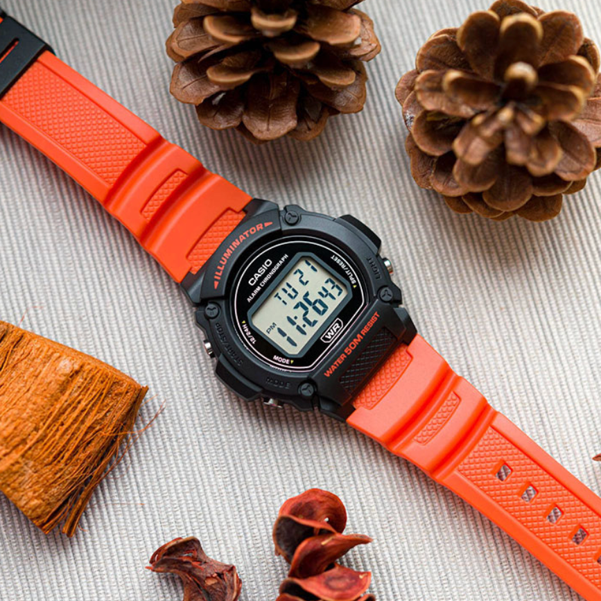 Digital watch with an orange strap on a textured surface with pine cones